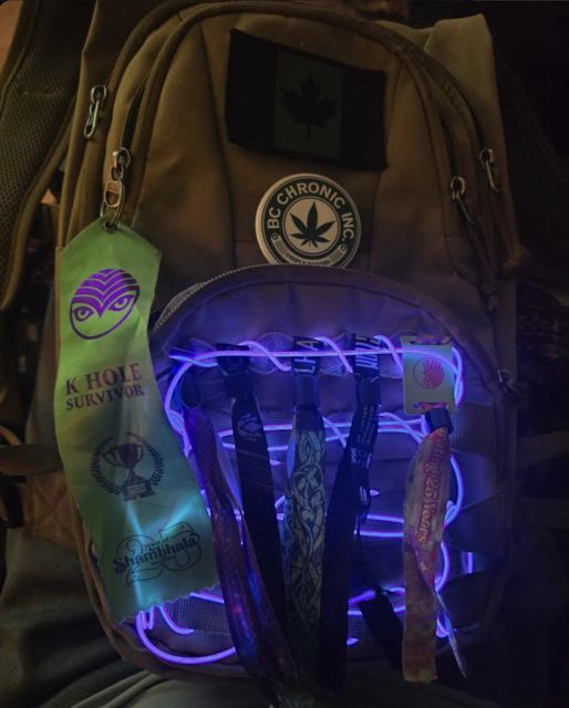 Tan backpack laced with glowing purple EL wire, hung with festival wristbands, a ribbon, and patches