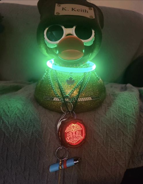 Gold glitter rubber duck in a black K. Keith beanie, round white sunglasses, a glowing green collar, chains and a Valhalla Fest badge